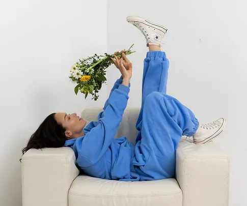 Person in a blue outfit lying on a white couch, holding a bouquet of flowers, wearing white sneakers.