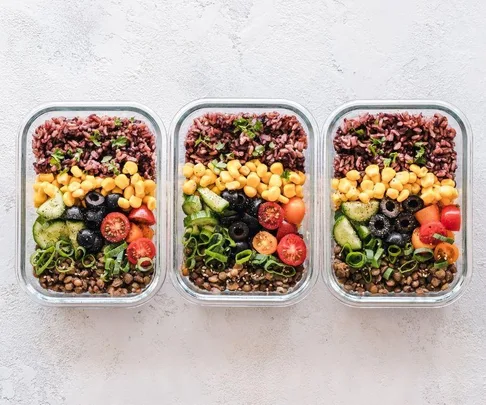 Three meal prep containers with rice, corn, black olives, cherry tomatoes, green onions, cucumbers, and parsley.