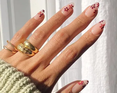 Hand with daisy nail art on pink nails, wearing gold rings, in natural light.