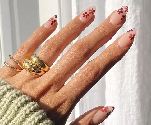 Hand with daisy nail art on pink nails, wearing gold rings, in natural light.