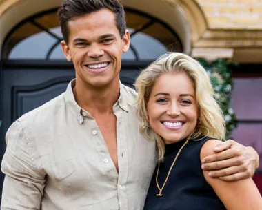 Two individuals smiling and embracing in front of a building with greenery.