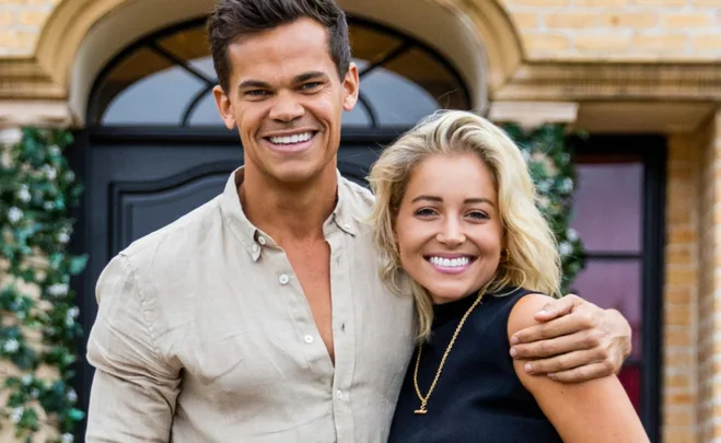 Two individuals smiling and embracing in front of a building with greenery.