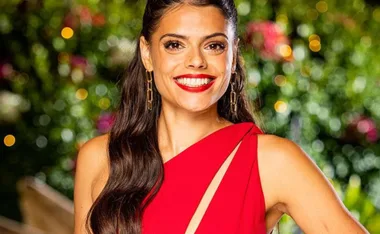 Smiling woman in a red dress with long hair and gold earrings, against a blurred garden background.