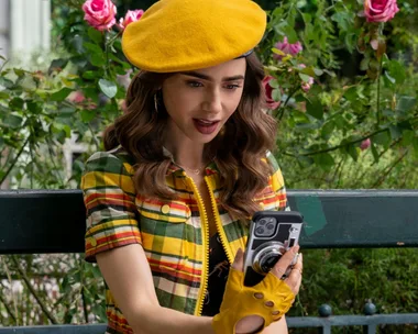 Emily in a yellow beret and plaid outfit takes a selfie on a bench surrounded by pink roses and greenery.