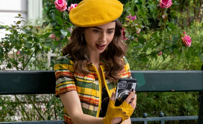 Emily in a yellow beret and plaid outfit takes a selfie on a bench surrounded by pink roses and greenery.