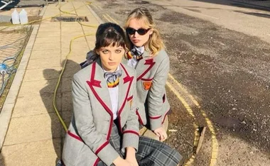 Two women in grey school uniforms with red trim, sitting on a sidewalk under bright sunlight.