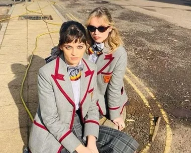 Two women in grey school uniforms with red trim, sitting on a sidewalk under bright sunlight.