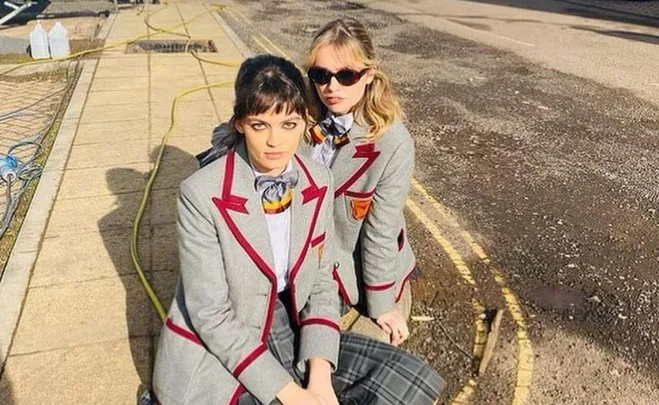 Two women in grey school uniforms with red trim, sitting on a sidewalk under bright sunlight.