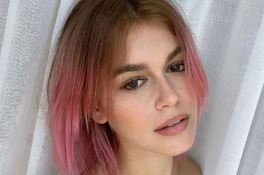 Woman with short, pink-highlighted hair against a white curtain backdrop.