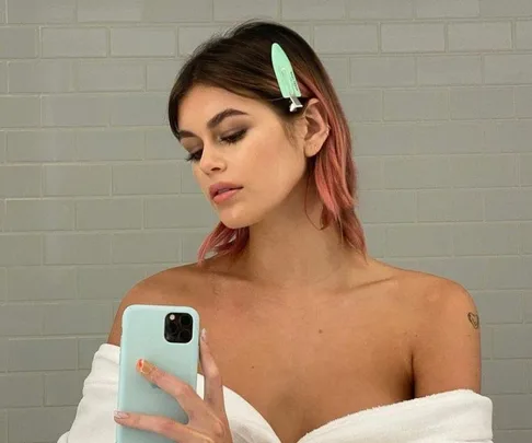 Woman with pink hair and green hair clip takes a selfie in a bathroom, wearing a white towel.