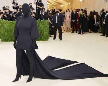 A person in an all-black, face-covering outfit with a long train on a red carpet at the Met Gala, surrounded by photographers.