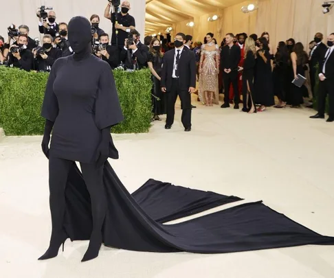 A person in an all-black, face-covering outfit with a long train on a red carpet at the Met Gala, surrounded by photographers.