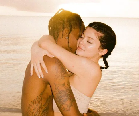 A couple embracing on a beach at sunset, with tattoos visible on the man's arm.