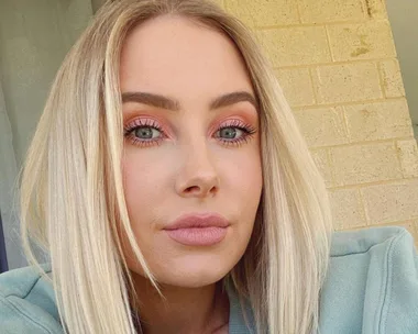 Blonde woman with striking eye makeup, wearing a casual green top, posing outdoors against a brick wall.