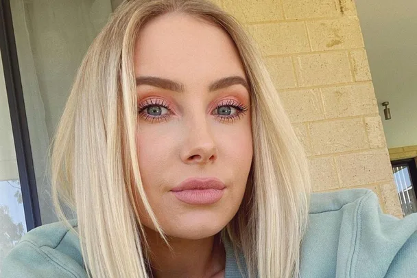 Blonde woman with striking eye makeup, wearing a casual green top, posing outdoors against a brick wall.