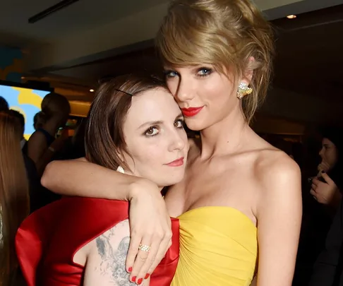 Two women posing closely at an event, one in a red dress and the other in a strapless yellow dress, smiling at the camera.