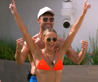 Man and woman in sunglasses celebrate enthusiastically outdoors, she wears an orange bikini, he wears a cap.