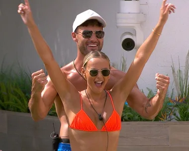 Man and woman in sunglasses celebrate enthusiastically outdoors, she wears an orange bikini, he wears a cap.