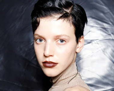 Model with short black hair and dark lipstick wearing a high-neck beige top, set against a textured silver backdrop.