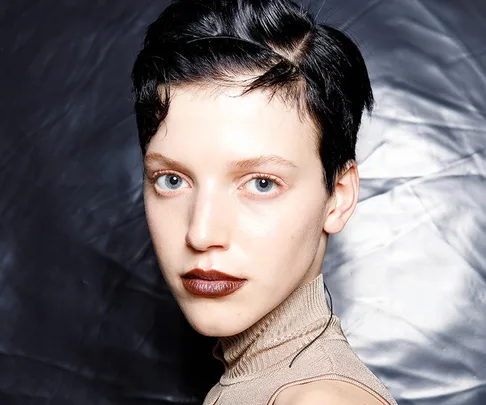 Model with short black hair and dark lipstick wearing a high-neck beige top, set against a textured silver backdrop.