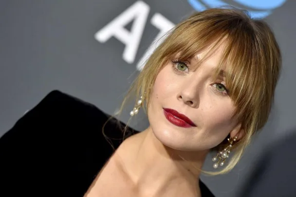 Woman with blonde hair and red lipstick in formal attire, posing on a gray carpet.