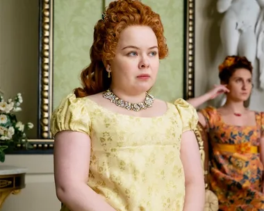 A woman in a yellow gown stands indoors, wearing a jeweled necklace, while another woman sits in the background.