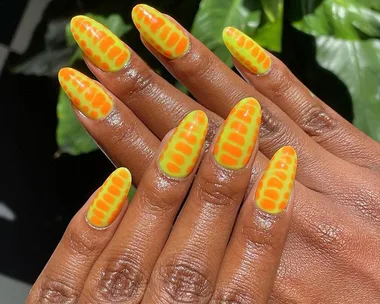 Bright orange and yellow nails with reptile skin pattern on dark skin, set against a leafy background.