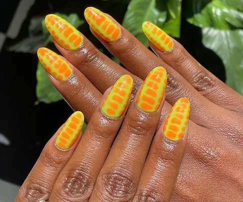 Bright orange and yellow nails with reptile skin pattern on dark skin, set against a leafy background.