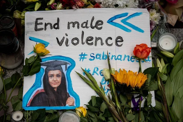 A tribute sign with "End male violence" and a photo of Sabina Nessa, surrounded by flowers and candles.