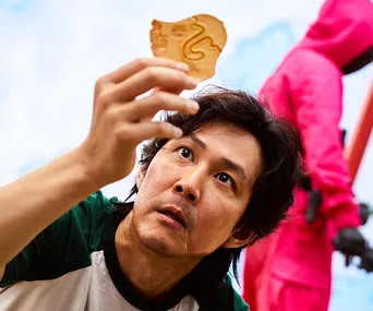 A man in a green shirt closely examines a honeycomb candy, with a guard in a pink suit in the background.