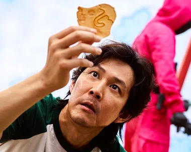 A man in a green shirt closely examines a honeycomb candy, with a guard in a pink suit in the background.