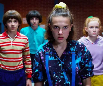 Characters from Stranger Things stand in colorful 80s clothes, led by a girl with a determined expression and ponytail.