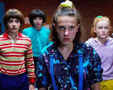 Characters from Stranger Things stand in colorful 80s clothes, led by a girl with a determined expression and ponytail.