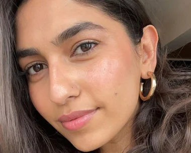 Close-up of a woman with long dark hair, wearing gold hoop earrings, in natural light.