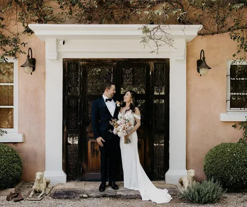 Bride and groom smiling in front of a vintage door, surrounded by greenery and statues.