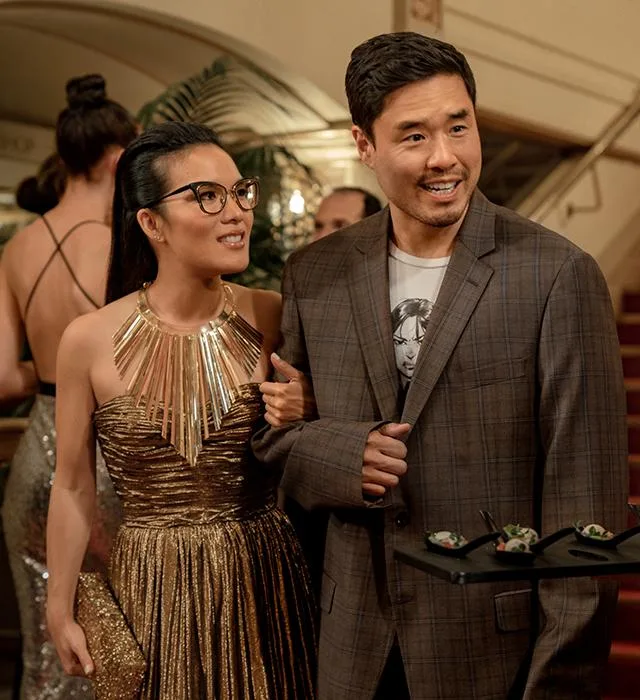 A couple dressed stylishly at a formal event, with the woman in a gold dress and glasses and the man in a plaid jacket.