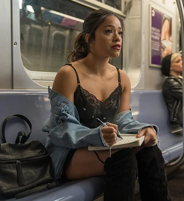 Young woman in a subway, writing in a notebook, wearing denim jacket and black lace top.