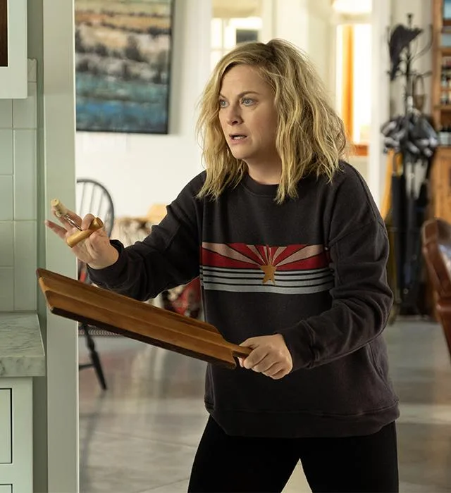 Woman in a dark sweater holding a cutting board and garlic in a bright kitchen.