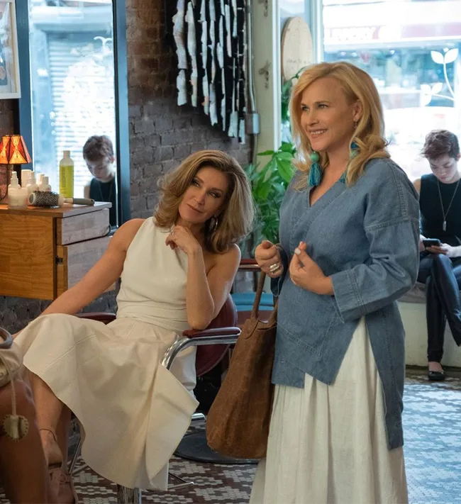 Two women are in a salon, one seated wearing a white dress, and the other standing in a blue top and white skirt, holding a bag.