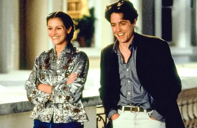 A man and woman smile while standing outside on a balcony at night, both casually dressed.