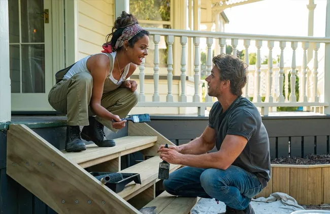 Two people painting steps, smiling at each other, on a porch. One is seated, and the other is crouching.