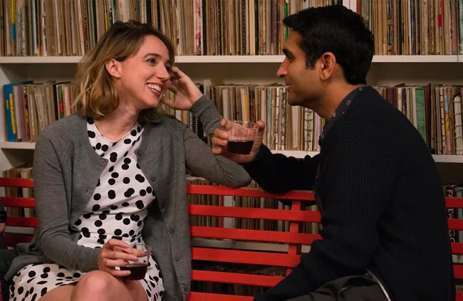 A woman in a polka dot dress and a man in a sweater sit on a red bench, smiling, holding drinks, with records behind them.