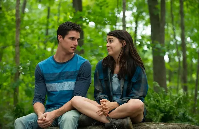 A young man and woman sit on a rock in a forest, engaged in a light conversation, surrounded by green trees.