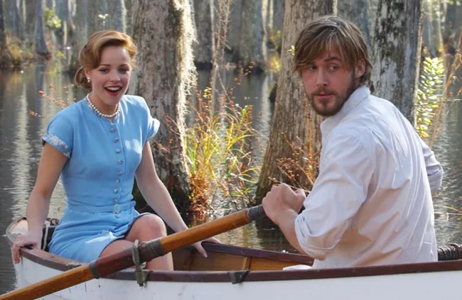 A couple in a rowboat on a serene lake, surrounded by trees. The woman wears a blue dress; the man, a white shirt.