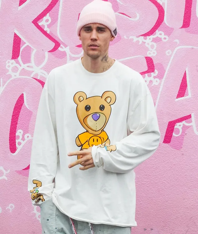 Man in pink beanie and teddy bear shirt poses in front of pink graffiti wall.