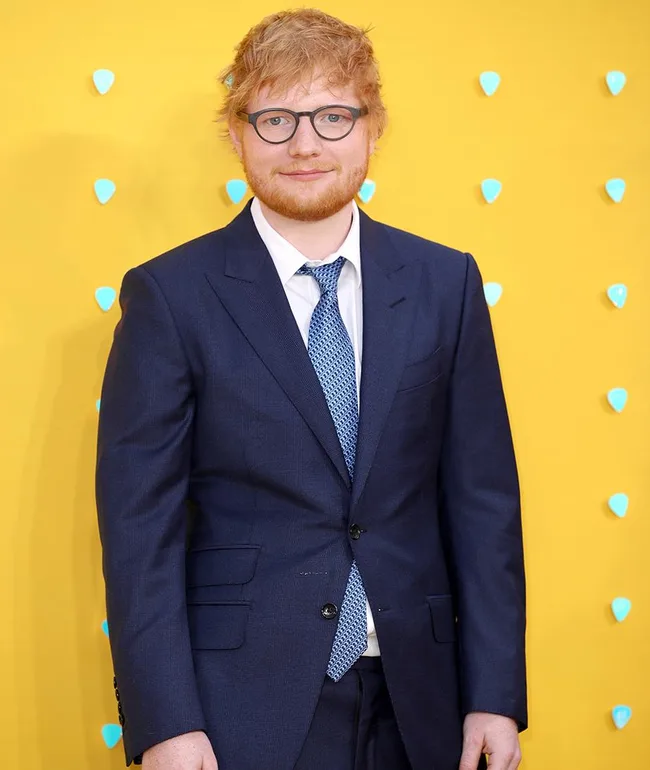 Man in a blue suit and glasses stands in front of a bright yellow background with blue dots.