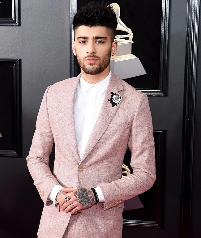 Man in a light pink suit with tattoos on hands, standing indoors with a Grammy award backdrop, looking at the camera.