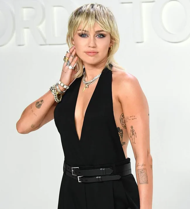 A person with short blonde hair and tattoos, wearing a black sleeveless outfit, poses at an event.