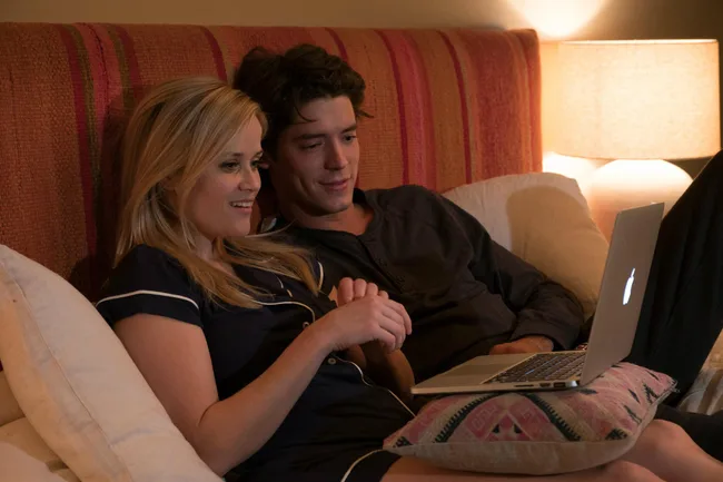 A woman and man, sitting on a bed, look at a MacBook screen with a lamp in the background.