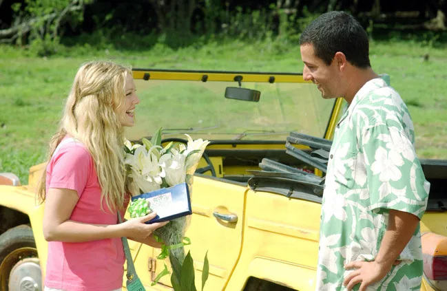A woman with blonde hair smiles, holding flowers and a gift, standing with a man in a green floral shirt near a yellow car.